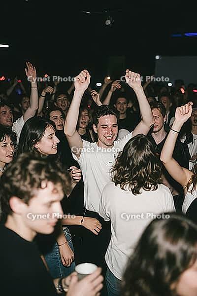 Ecstatic youth celebrating togetherness on a crowded nightclub dancefloor, joyful party culture embodying social cohesion and vitality, a smiling young man in a white shirt raises both arms into the air as a profound expression of liberation, uninhibited  [IBR125446466]