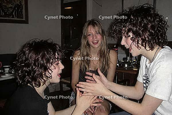 Three young friends enjoying playful hand games during an evening gathering, sharing authentic joy, laughter and emotional connection, dynamic gesture coordination between two companions clapping hands while a smiling girl observes with cheerful amusement [IBR125446471]