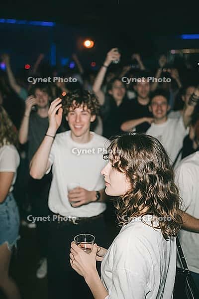 Vibrant nightlife youth culture party in packed club venue with dancing young adults holding drinks, collective euphoria and nocturnal social bonding amidst atmospheric stage lights, young woman in contemplative profile holding beverage cup surrounded by  [IBR125446481]