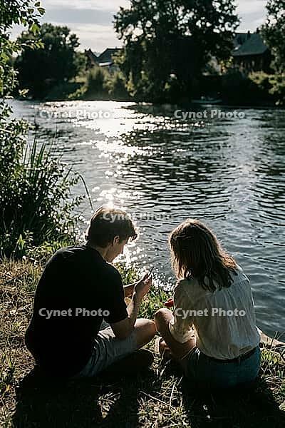 Youthful romance, tranquil riverbank intimacy, sparkling water reflections of summer light, two young people sit close together at the grassy river shore, backlit silhouettes immersed in serene quietude, shimmering sun glitter on flowing ripples, the flow [IBR125446484]