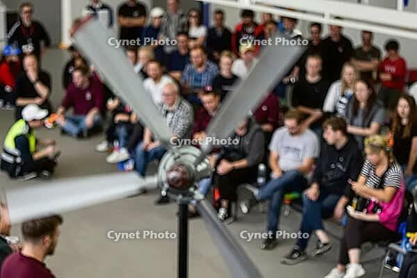 Engineering students attend aerodynamic wind turbine demonstration in technical university lecture hall, focus on mechanical rotor blades, blurred audience of young academics observing technological presentation, conceptual synergy between renewable energ [IBR125446493]