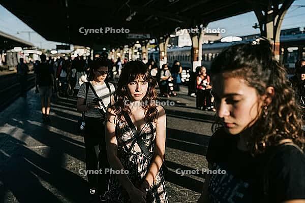 Urban isolation amidst crowded public transit, an introspective woman stands frozen on a sunlit train platform, deep contemplative vulnerability contrasted against moving commuter streams, rhythmic shadow patterns across concrete surfaces embody the psych [IBR125446495]