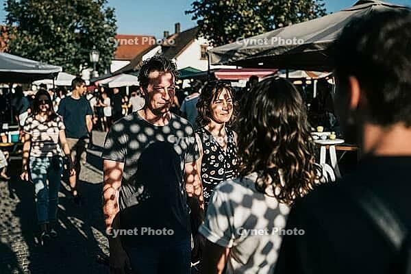Outdoor market gathering in warm sunlight, visitors chatting on cobblestone pavement under canvas canopies, polka dot shadows filter through perforated shades creating geometric patterns across human faces and casual summer clothing, intense eye contact b [IBR125446503]
