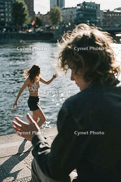 Youthful vitality meets urban waterside escapism, blurred figures engaging in spontaneous gestures while sparkling sunlight reflects off ripples, a dancing woman in light attire balances gracefully along the stone riverbank embankment, shallow depth of fi [IBR125446510]