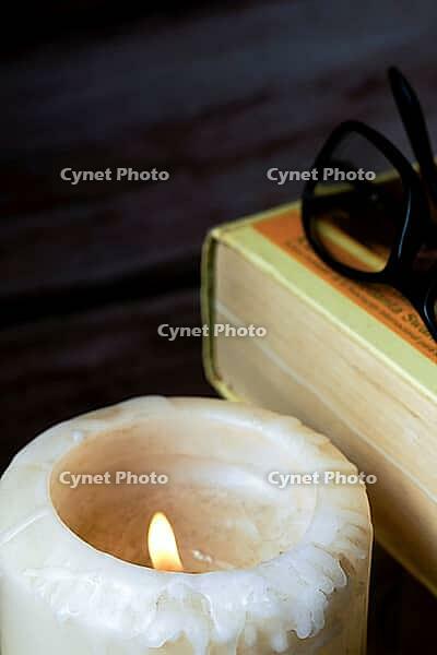 Burning candle providing warm light for a vintage book and reading glasses, symbolizing education, knowledge, and quiet study [IBR125448817]