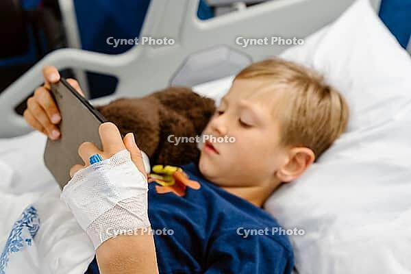 Sick boy lying in a hospital bed, playing on his phone with a teddy bear beside him, Portugal [IBR125448829]