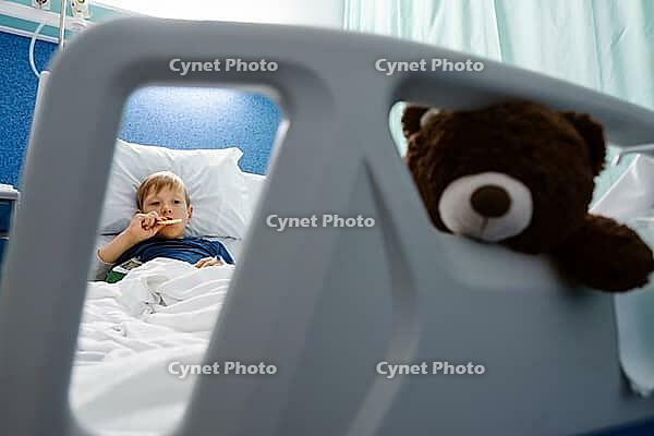A young boy resting and eating in a hospital bed while holding a teddy bear [IBR125448843]