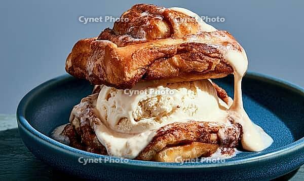 Sweet cinnamon roll piled high with vanilla ice cream. Creamy sauce drizzles down sides. Served on dark blue plate, AI generated [IBR125448878]