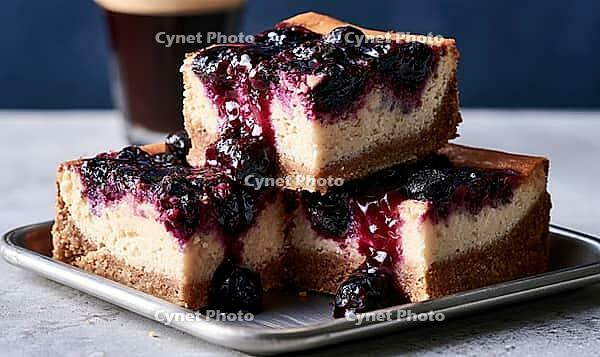 Three blueberry cheesecake slices stacked on a metal tray. Dark blueberries glisten on top of creamy cake. Warm lighting highlights texture, AI generated [IBR125448883]