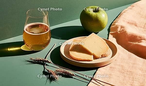 Glass of beer beside green apple and cheese slices on wooden plate. Warm sunlight casts soft shadows on green and pink surface, AI generated [IBR125448886]