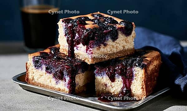 Three square blueberry cheesecake pieces on metal tray. Berry sauce drips over golden crust. Dark coffee mug sits behind, AI generated [IBR125448893]
