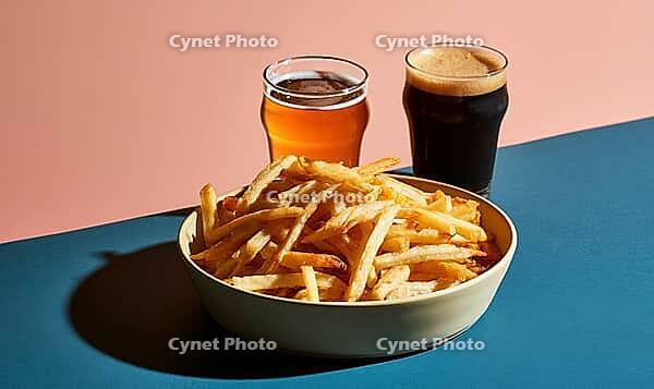 Bowl of golden french fries sits on blue surface. Two glasses of beer stand behind. One glass contains amber beer, other dark beer, AI generated [IBR125448895]