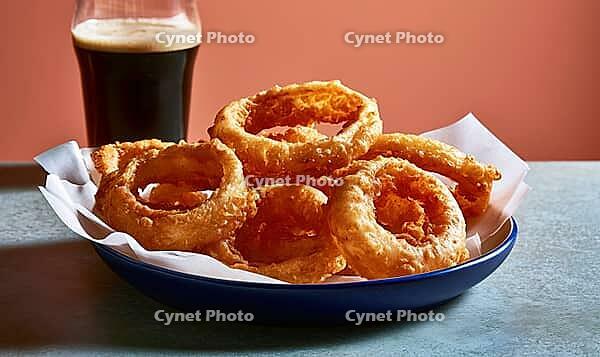 Crispy fried onion rings sit in blue dish. Glass of dark beer sits beside. Food rests on white paper. Background is warm orange wall, AI generated [IBR125448897]