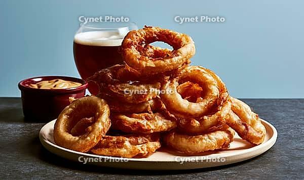 Golden, crispy onion rings piled on a white plate. Beside them, a glass of dark beer and a small dipping sauce bowl, AI generated [IBR125448903]
