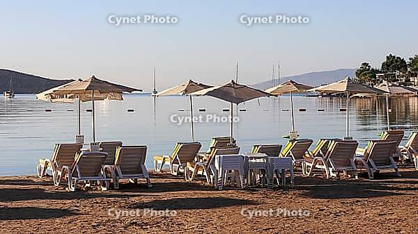 Beach chairs and umbrellas line a sandy shore, overlooking a calm sea with boats and distant hills under a clear sky. [IBR125462056]