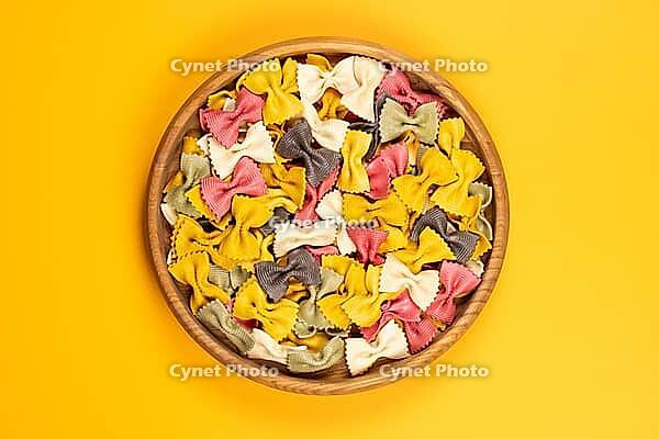 Farfalle pasta of various colors filling a wooden bowl, presented as dry ingredients on a vibrant yellow background and offering a cheerful culinary theme [IBR125462086]