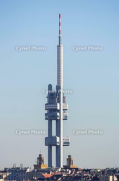 Prague, Czech Republic - February 15, 2019: The Zizkov Television Tower rises above the rooftops of the Zizkov district against a clear blue sky, with church towers and residential buildings visible at the base [IBR125462089]