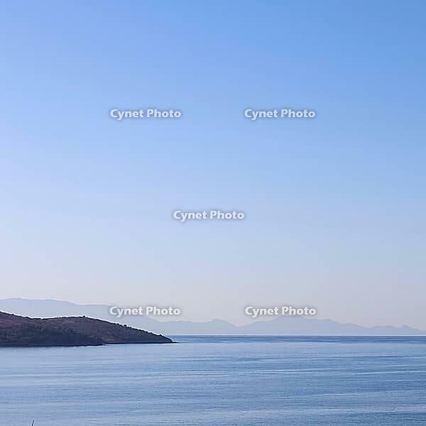 A tranquil blue seascape with a dark coastline in the foreground, and hazy distant mountains fading into a clear, gradient sky. [IBR125462096]