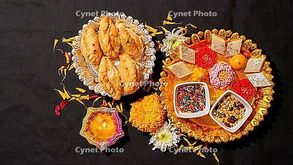 A top-down view of traditional Indian sweets on silver and gold platters, a burning clay diya, marigold flowers, and bowls of sprinkles on a black background [IBR125462097]
