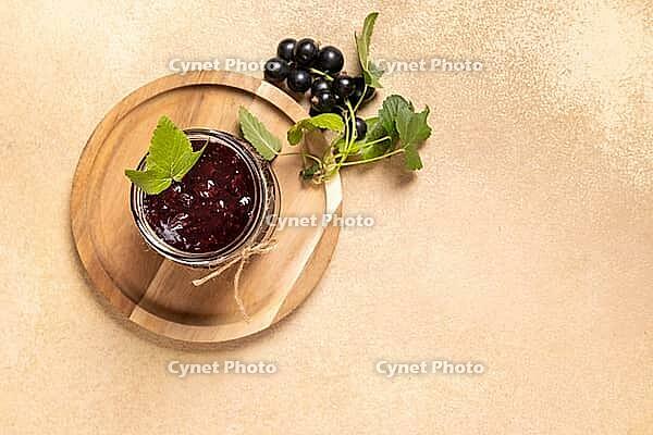 Black currant jam, prepared at home, filling a glass jar on a wooden serving board, featuring fresh berries and green leaves, representing sweet fruit preservation and delicious dessert spread [IBR125462103]