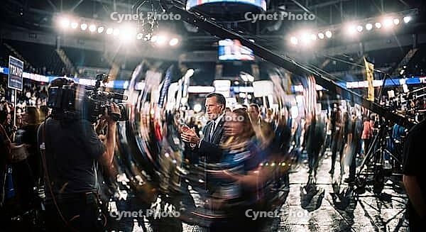 Political campaign rally inside a crowded sports arena with broadcast camera equipment, television journalists and a clapping politician in the center, media spectacle and public relations staging within modern democratic election processes, large televis [IBR125472246]