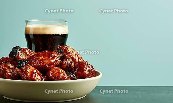 Bowl of glazed chicken wings with beer glass behind. Wings sit on white plate against teal background. Food photo with warm lighting, AI generated [IBR125472268]