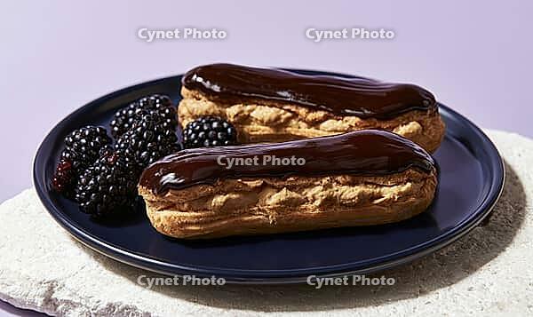 Two chocolate eclairs sit on dark plate. Blackberries beside pastries add color contrast. Plated dessert ready for serving or photography, AI generated [IBR125472290]