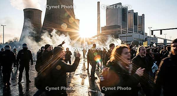 Climate justice activism confronting fossil fuel infrastructure, riot police lines facing shouting youth protesters amid drifting smoke and blinding lens flare, massive cooling towers and industrial smokestacks towering above turbulent civil disobedience  [IBR125472293]