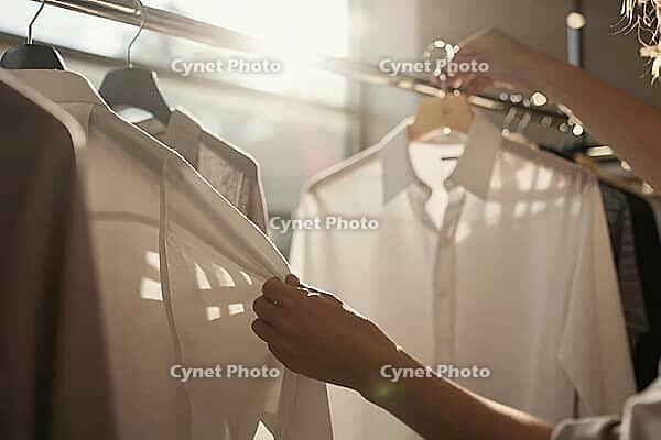 Woven cotton shirts hang on a polished metal garment rack in bright sunlight as hands gently touch soft textile fabrics during clothing selection, tactile sensory examination of high quality tailored garments underscores mindful consumer choices and slow  [IBR125472299]