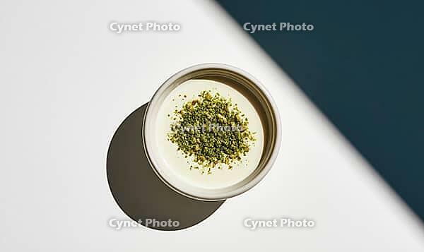 Top-down view of a white bowl with creamy yogurt. Yogurt topped with crushed green pistachios. Bowl placed on white and dark blue surface, AI generated [IBR125472301]
