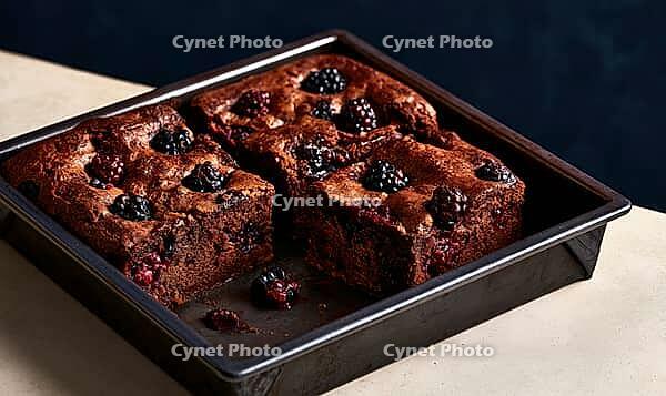 Freshly baked chocolate brownies with blackberries. Served in metal baking pan. Rich, moist texture visible. Dark background enhances warm tones, AI generated [IBR125472305]