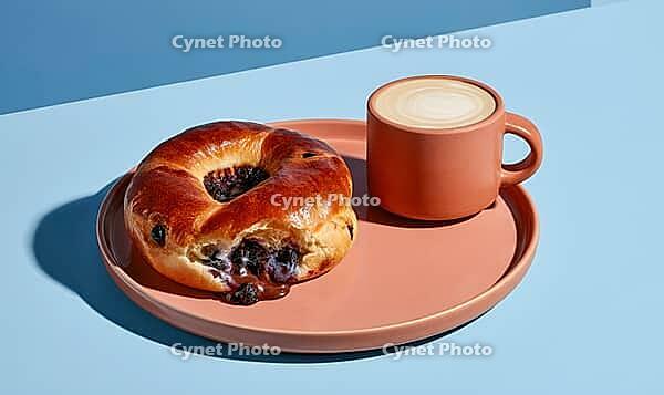 Golden-brown blueberry-filled bagel on terracotta plate. Next to cup of frothy coffee. Set against clean blue background, AI generated [IBR125472306]