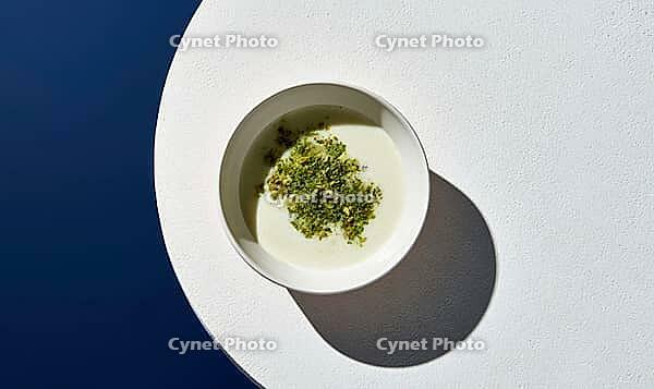 White bowl holds creamy soup with fresh green herbs. Bowl sits on white circular plate. Deep blue background contrasts with light tones, AI generated [IBR125472324]