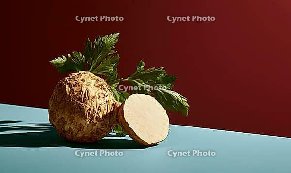 Celeriac root with fresh green leaves on blue surface. One half sliced, showing pale interior. Deep red background contrasts with cool tones, AI generated [IBR125472333]