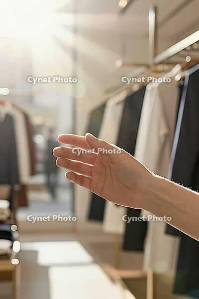 Delicate fingers reaching toward textiles along minimalist boutique racks, sensory consumer exploration within bright retail spaces, graceful hand movement symbolizing tactile desires in contemporary commerce, warm morning daylight filtering through store [IBR125472334]