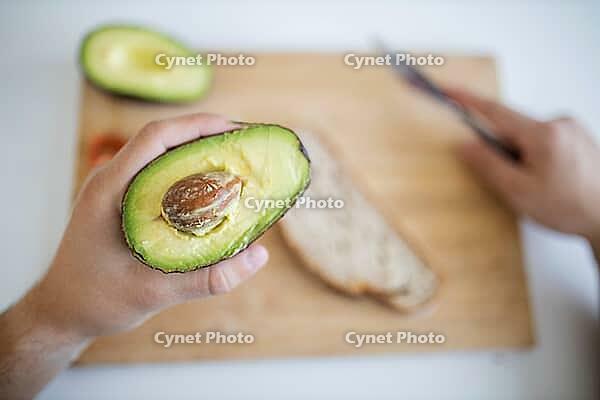 Male hand holding half avocado over blurry wooden cutting board with bread and tomatoes. Man at white table preparing avocado toast. Healthy meal preparation [IBR125492330]