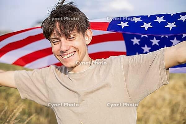 Happy and cheerful teenage boy with arms spread wide waving American flag in a field outside the city. [IBR125492332]