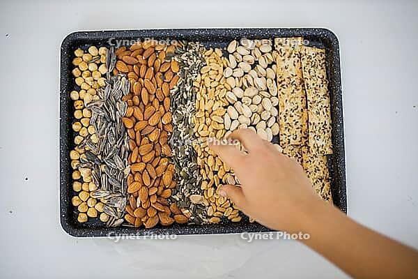 Female hand picking peanuts from black tray made of chickpeas, almonds, pistachios and multiple sawn crisp bread. Fresh crispy bread, seeds and nuts from above. Healthy snacks [IBR125492340]