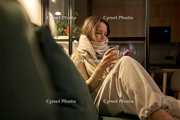 Female seated on sofa wrapped in thick scarf holding a glass of water and smartphone, viewed in warm indoor lighting with a relaxed front angle and visible living room elements [IBR125492347]