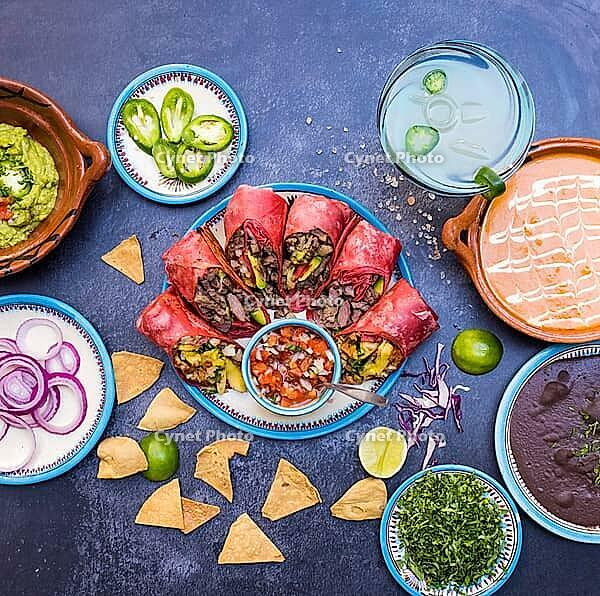 Top view of six delicious burritos, pico de gallo and traditional dishes on dark grunge surface. Tortilla chips, sliced peppers, fried beans and more on plates. Authentic Mexican cuisine [IBR125492351]