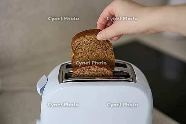 Handhalten Single Brown Bread Slice oben Weiß Zwei Slice Toaster auf Küchenarbeitsplatte in der Nähe von Black Electric Kochplatten im Innenbereich Schließen Sie nach oben Angehobene Slice bereit für Einlegearbeiten [IBR125492365]