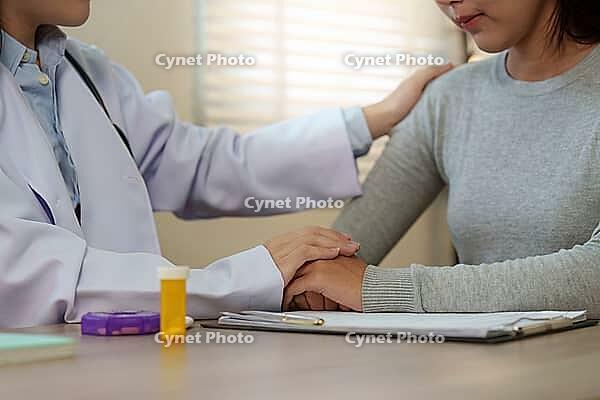 A healthcare professional in a white coat places a reassuring hand on the shoulder of a sad young Asian woman, offering support and empathy during a clinic visit [IBR125492374]