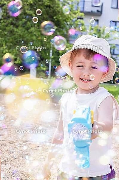 A cheerful toddler boy wearing a bucket hat laughs while shooting soap bubbles from a blue toy gun on a bright, sunny day outdoors [IBR125492375]