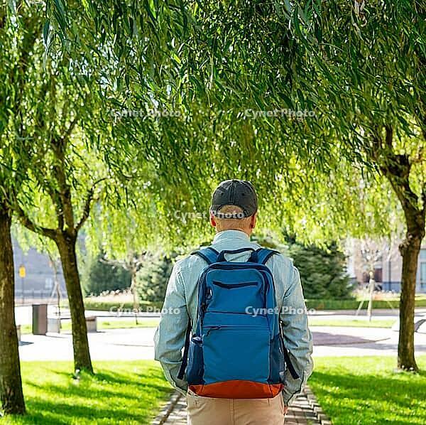 Man carrying backpack under willow tree in municipal park. Travelers explore nature and urban landscape. Concept of travel, adventure and leisure in summer sunshine. Rear view [IBR125492383]