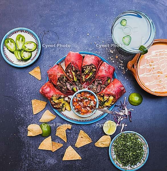 Top view of delicious burritos, pico de gallo, and more traditional food on dark grunge surface. Tortilla chips, sliced peppers, sauce and more on plates and clay pots. Authentic Mexican cuisine [IBR125492388]