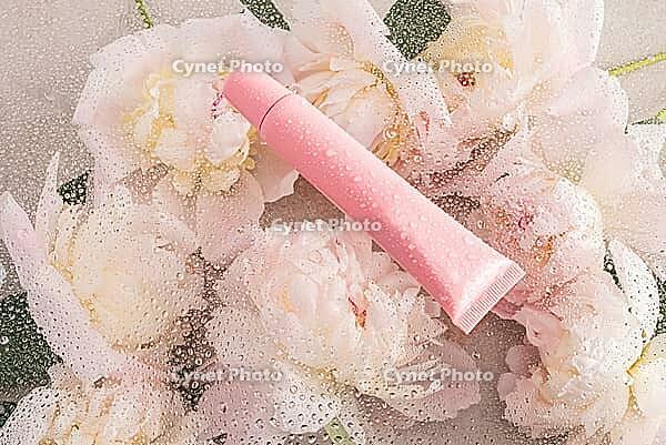 A pink plastic tube with a moisturizing balm, lip gloss, lip care mask lies on a chic background with delicate peonies and drops of water. empty package [IBR125492403]