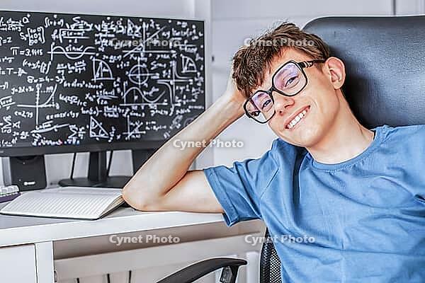 Cute teenage student in glasses sitting at a computer studying online, looking at the camera and smiling contentedly. [IBR125492406]