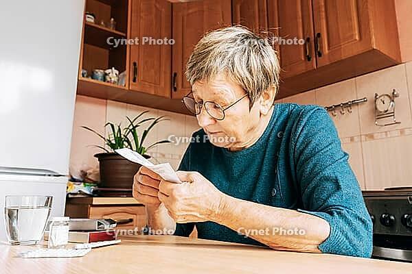 An elderly woman reads pill instructions in kitchen setting. Health maintenance, healthy aging, vitamin support, home treatment [IBR125492416]