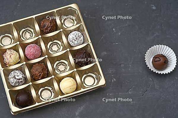 Chocolate assortment presented in square golden box on dark surface. Chocolate sweets with varied shapes and coatings, elegant pattern. premium dessert concept, holiday gift, indulgence background [IBR125492417]