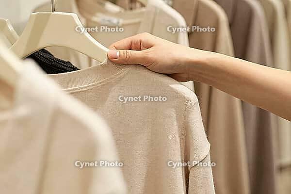 Tactile fashion retail experience with neutral beige knitwear on hangers, a customer hand touches soft wool textiles in a quiet luxury boutique, delicate fingers gently examining the knit structure of an understated beige sweater on a minimalist hanger co [IBR125502124]
